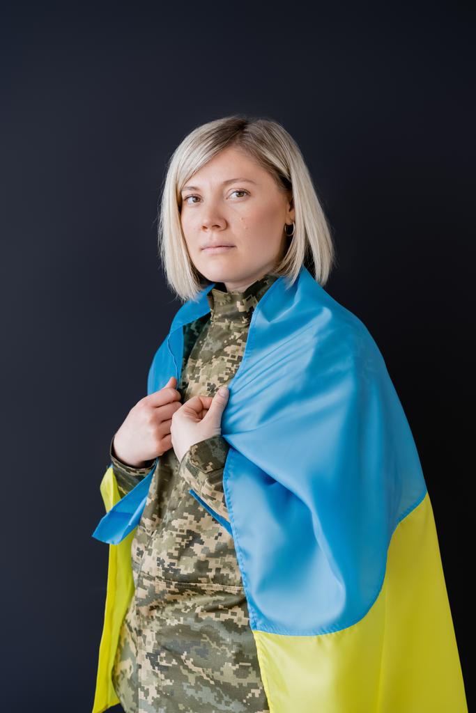 Omuzlarında Ukrayna bayrağı olan askeri üniformalı bir kadın siyah kameraya bakıyor. - Fotoğraf, Görsel