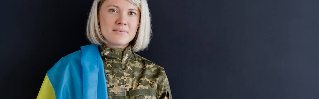 Ukrayna bayrağı taşıyan sarışın asker kadın siyah pankartta izole edilmiş kameraya bakıyor. - Fotoğraf, Görsel