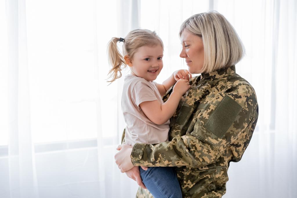 γυναίκα με στρατιωτική στολή που κρατά ευτυχισμένη κόρη στο σπίτι - Φωτογραφία, εικόνα