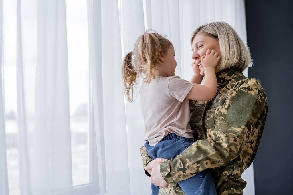 niña con coletas tocando la cara de mamá sonriente en uniforme militar cerca de la ventana - Foto, imagen