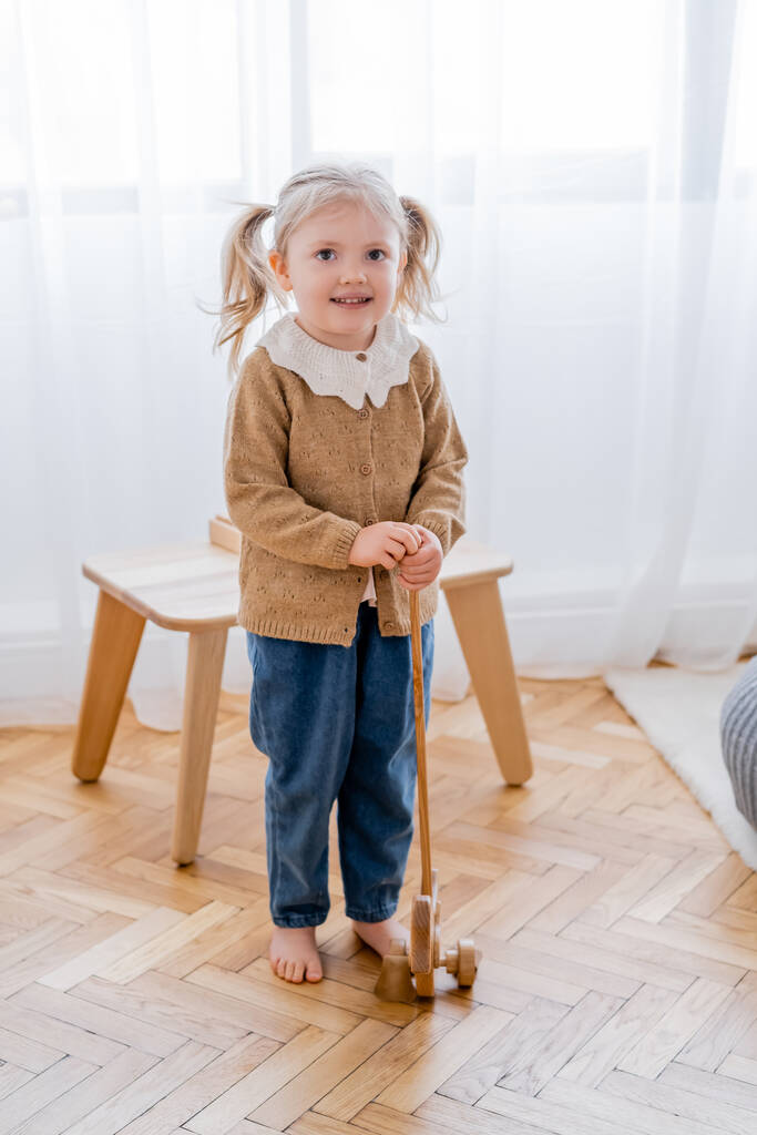 full length view of barefoot girl with wooden toy smiling at camera - Photo, Image