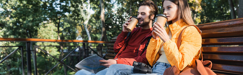 Positiivinen matkustaja tilalla kartta ja kahvia mennä lähelle tyttöystävä penkillä ulkona, banneri  - Valokuva, kuva