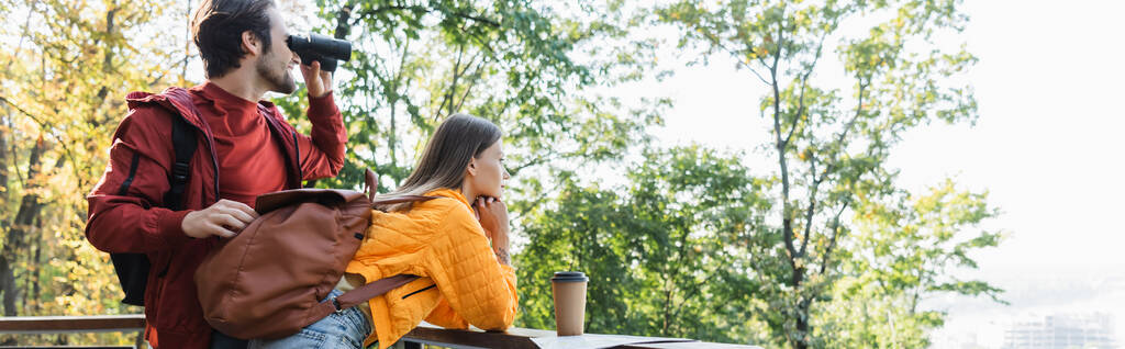 Vue latérale du touriste regardant à travers des jumelles près de petite amie avec sac à dos, café et carte en plein air, bannière  - Photo, image