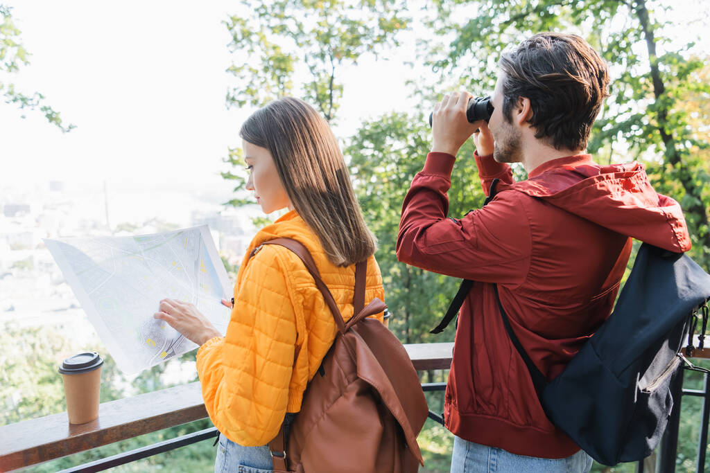 Side view of young woman looking at map near coffee to go and boyfriend with binoculars outdoors  - Photo, Image
