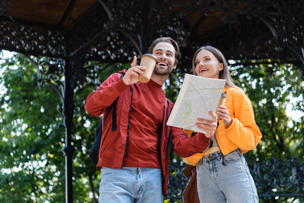 笑顔の観光客が紙コップと地図を保持しながら、指で指している間の低角度ビュー屋外のガールフレンド  - 写真・画像