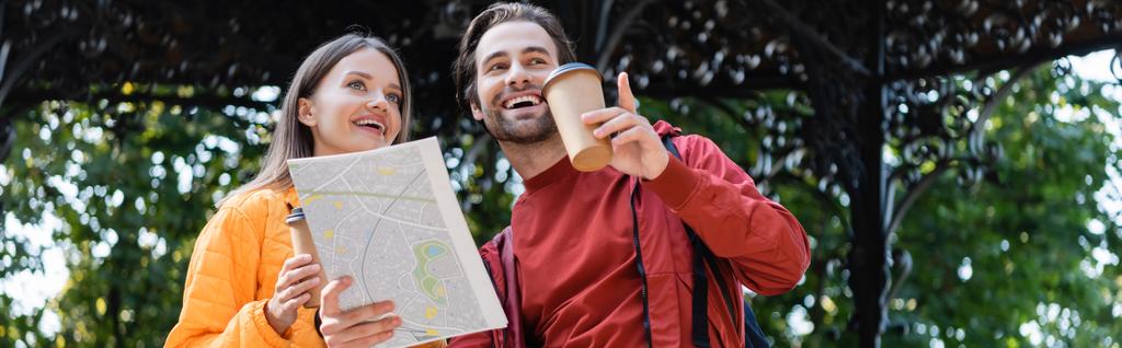 Низький кут зору веселий чоловік тримає карту і вказує пальцем біля дівчини з кавою, щоб піти на міську вулицю, банер
  - Фото, зображення