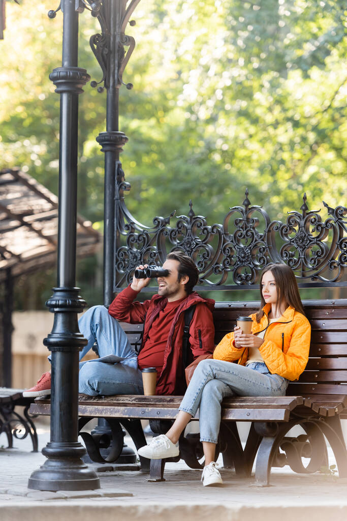 Glücklicher Tourist schaut durch Ferngläser in der Nähe Freundin mit Kaffee zu gehen sitzen auf Bank im Freien  - Foto, Bild
