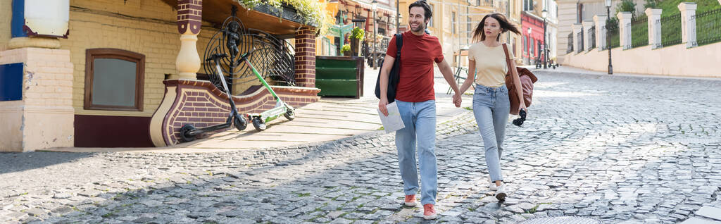 Пара путешественников с картой и биноклем держатся за руки во время прогулки по городской улице, баннер  - Фото, изображение