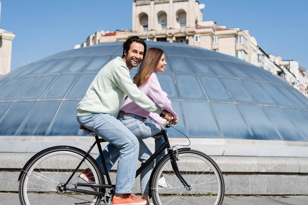 Sonriente hombre mirando a la cámara mientras pedalea con su novia en la calle urbana  - Foto, imagen