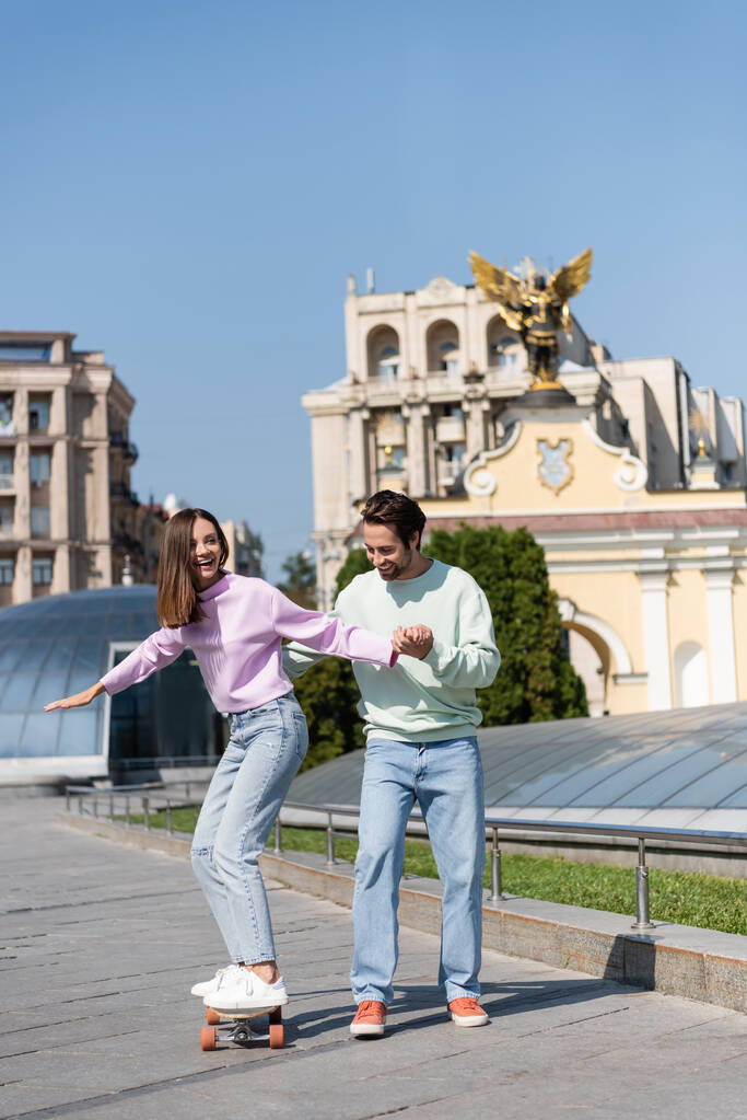 Позитивная пара в толстовках и джинсах, катающаяся на копеечной доске на городской улице  - Фото, изображение