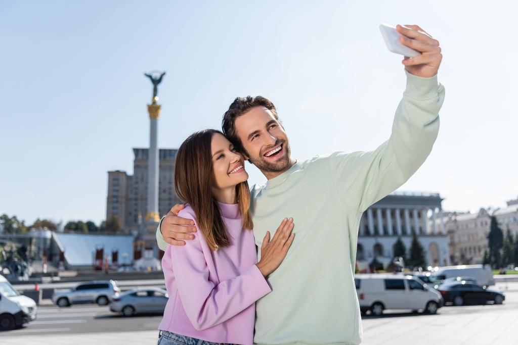 KYIV, UKRAINE - SZEPTEMBER 1, 2021: Vidám pár ölelés és selfie okostelefonon a Függetlenség terén - Fotó, kép