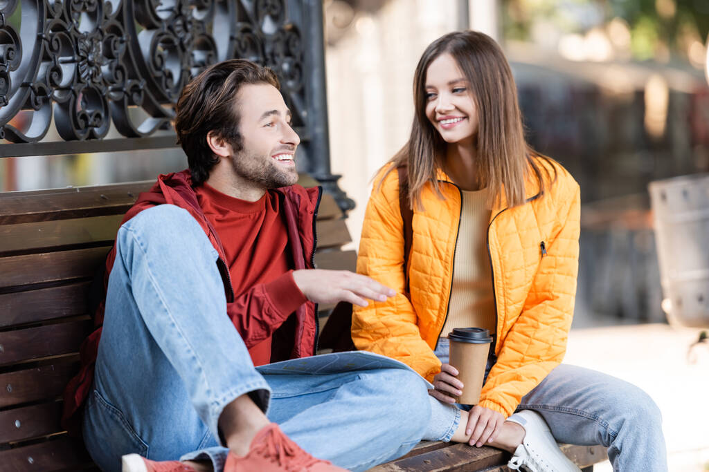 Улыбающийся мужчина разговаривает с девушкой с кофе, чтобы пойти на скамейку запасных на городской улице  - Фото, изображение