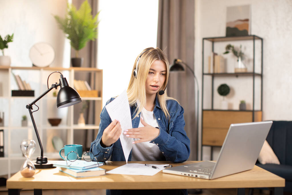 Femme blonde en casque et vêtements décontractés ayant un chat vidéo sur ordinateur portable, travaillant à la maison ou au bureau lumineux - Photo, image