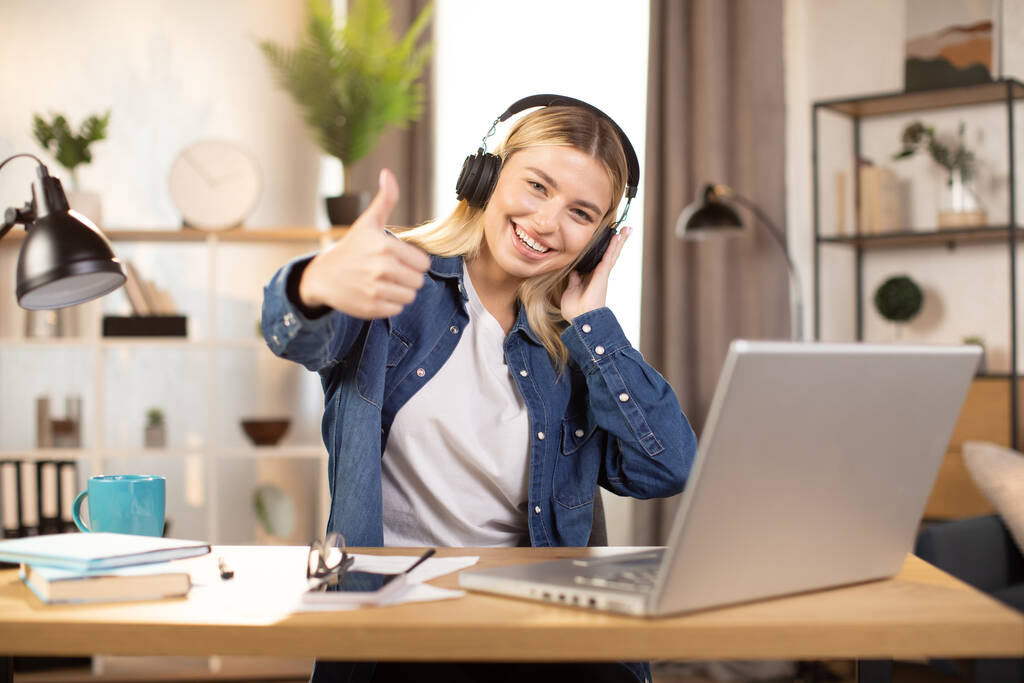 woman wearing headphones, sitting at the table at modern apartment, and showing victory sign gesture - Фото, изображение