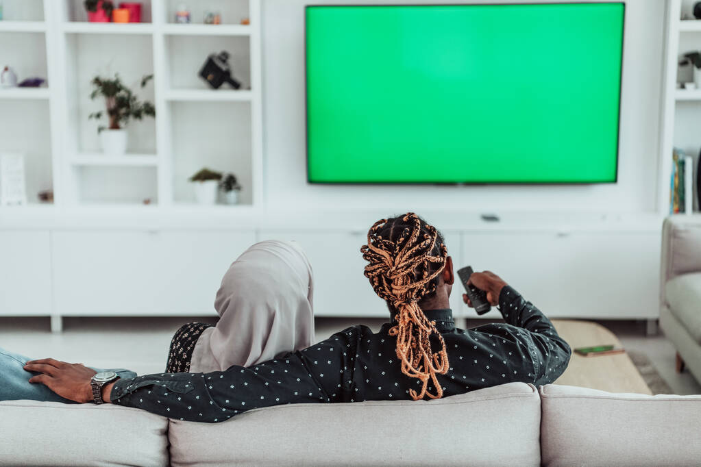 ソファに座っているアフリカのカップルテレビを一緒に見るクロマ緑の画面イスラム教のヒジャブを身に着けている女性服 - 写真・画像