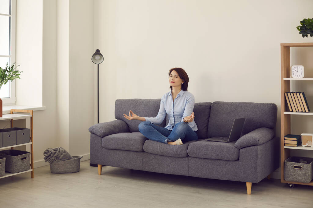Επιχειρηματίας κυρία κάνει ασκήσεις γιόγκα για την ανακούφιση από το άγχος, ενώ κάθεται στον καναπέ στη θέση του λωτού. - Φωτογραφία, εικόνα