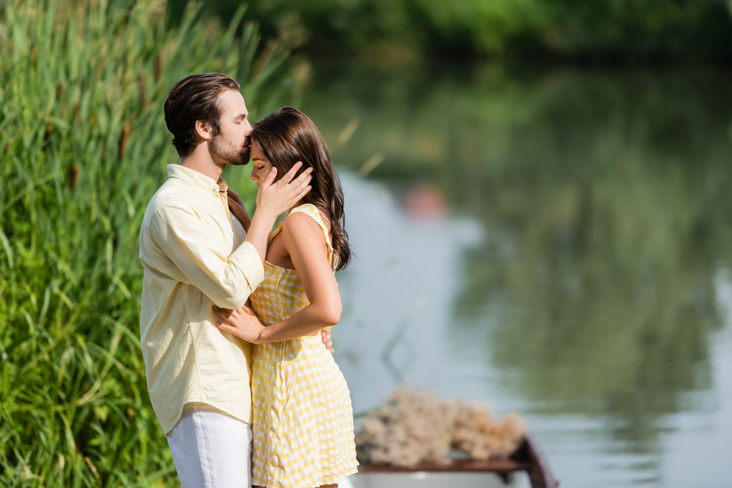 πλαϊνή άποψη του γενειοφόρου άνδρα φιλιά μέτωπο της νεαρής γυναίκας στο φόρεμα κοντά στη λίμνη - Φωτογραφία, εικόνα