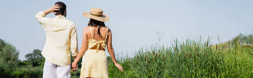 вид на молодоженов в летней одежде, держащихся за руки и стоящих у озера, баннер - Фото, изображение