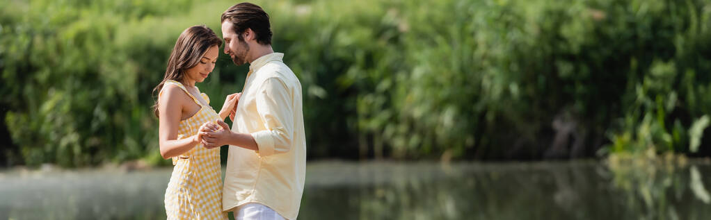 счастливая молодая пара в летней одежде, держась за руки у озера, баннер - Фото, изображение