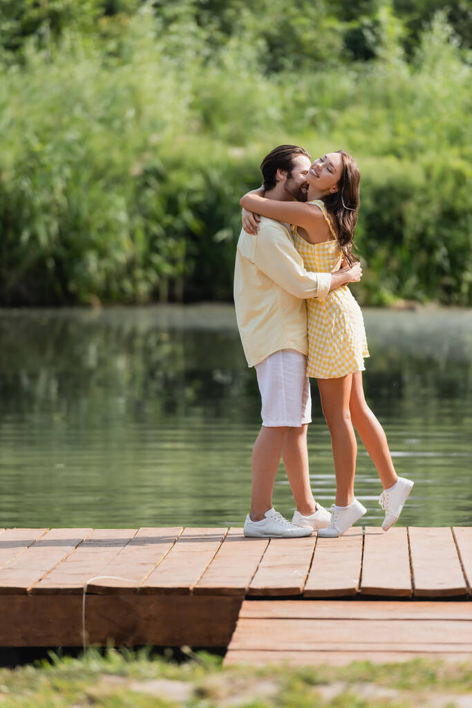 full length of joyful young couple in summer clothes hugging on pier near lake - Photo, Image