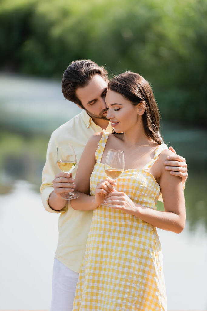 romántico joven pareja en sonriendo y sosteniendo vasos con vino cerca del lago - Foto, imagen