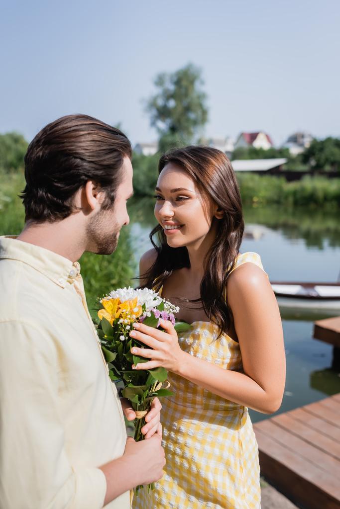 Sakallı bir adam elinde çiçek buketi, göl kenarında giyinmiş neşeli bir kadının yanında.  - Fotoğraf, Görsel