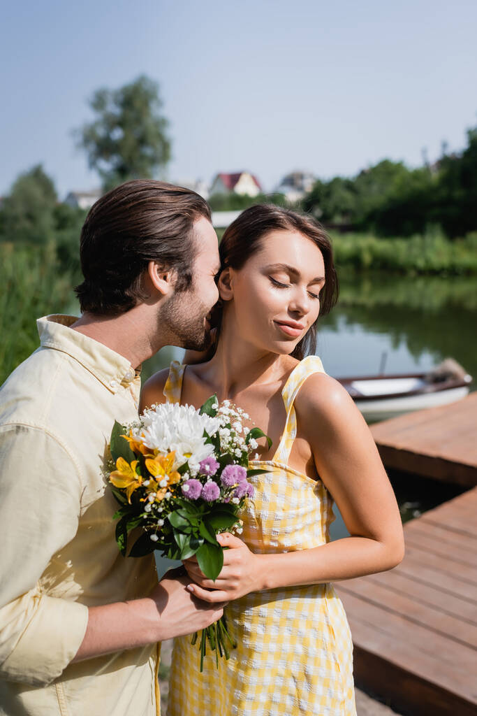 homem barbudo dando buquê de flores e beijando mulher perto do lago  - Foto, Imagem