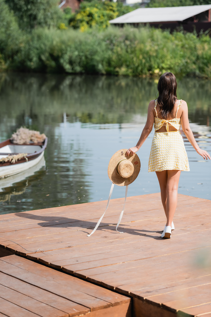 Yazlık elbiseli genç bir kadın göl kenarında hasır şapka tutuyor. - Fotoğraf, Görsel