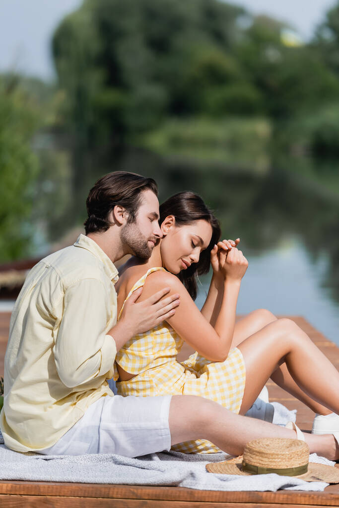 hombre abrazando sensual joven mujer mientras está sentado en el muelle cerca del lago - Foto, Imagen