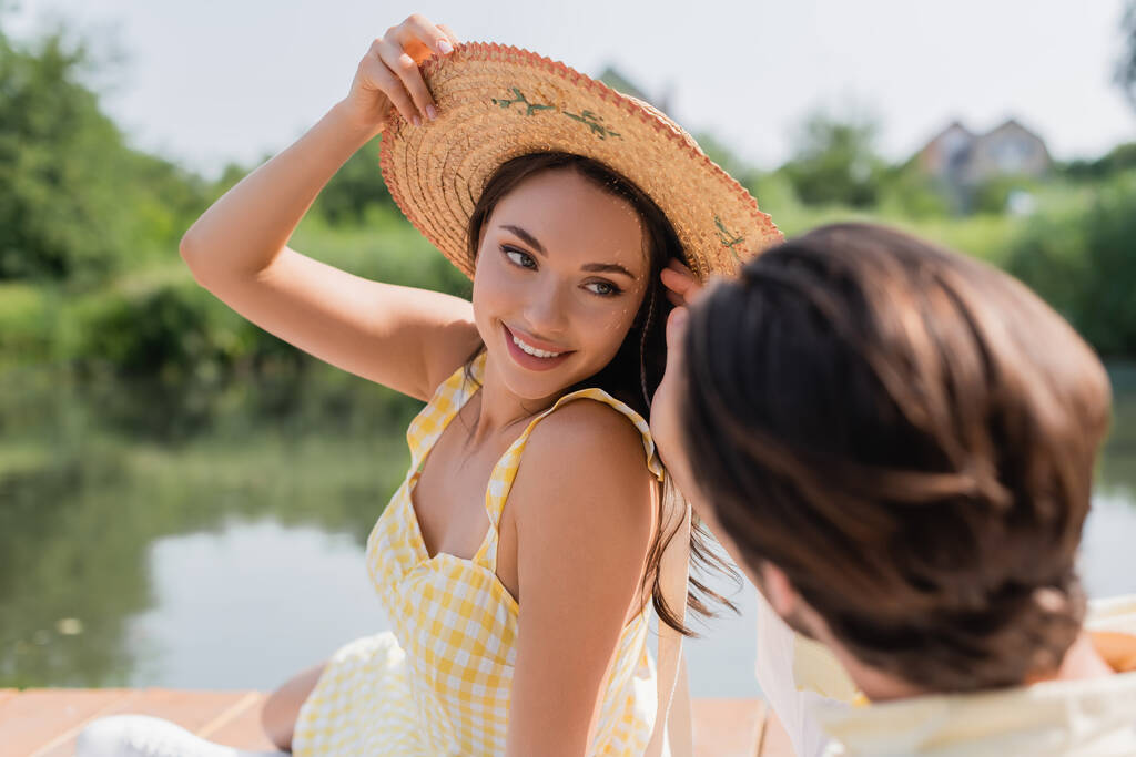 homme flou regardant femme souriante ajuster chapeau de paille tandis que près de la rivière - Photo, image