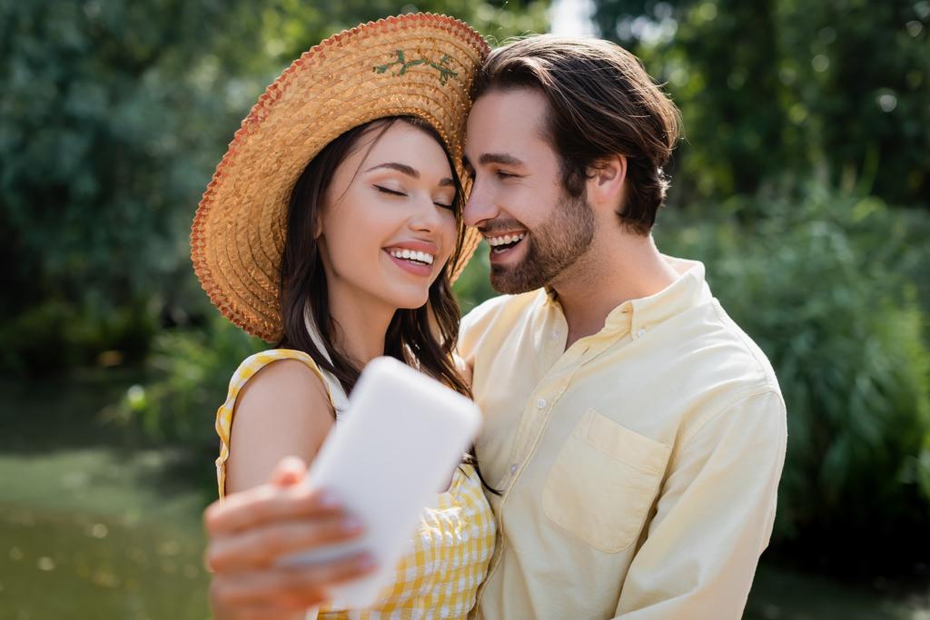 gelukkig jong vrouw in stro hoed het nemen van selfie met vrolijke vriend buiten - Foto, afbeelding