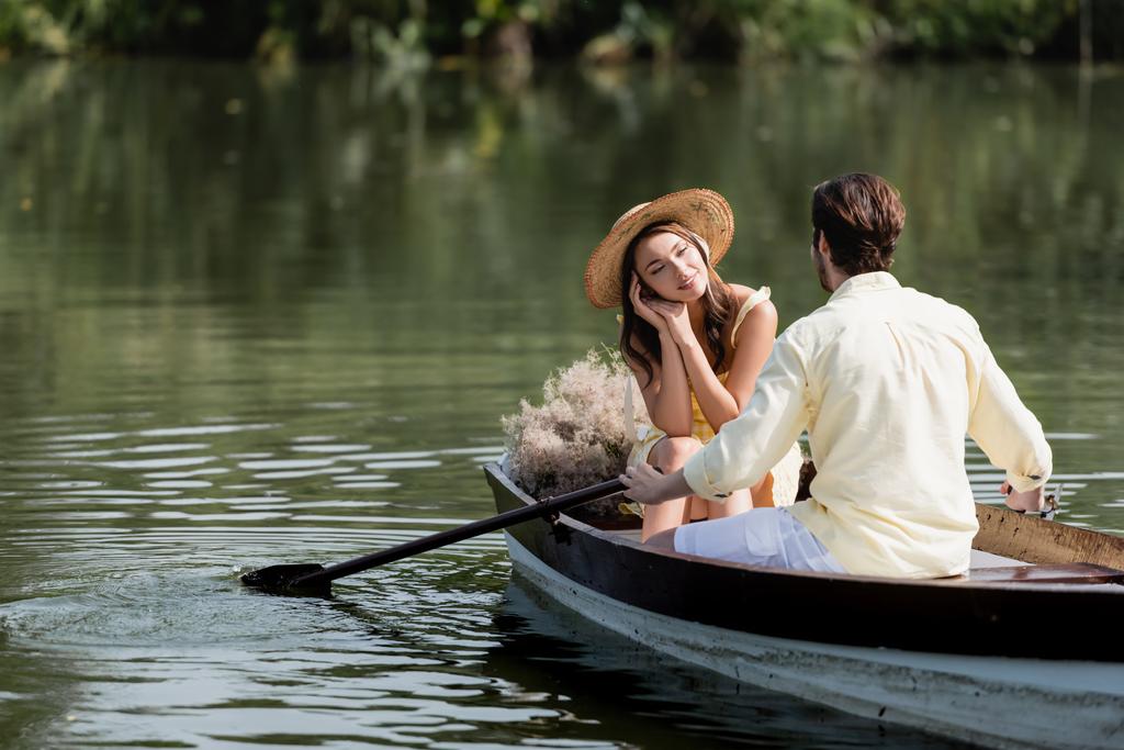 dromerige vrouw in stro hoed kijken naar romantisch vriendje tijdens boottocht - Foto, afbeelding