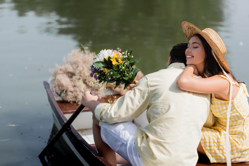 Hasır şapkalı mutlu genç bir kadın elinde bir buket çiçek ve domuz gezisi sırasında erkek arkadaşına sarılıyor. - Fotoğraf, Görsel