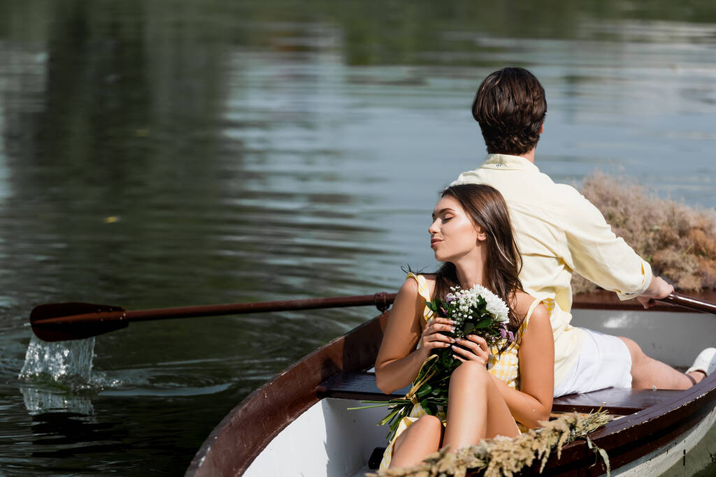 Romantik tekne gezisi sırasında elinde çiçeklerle erkeklerin sırtına yaslanan genç bir kadın.  - Fotoğraf, Görsel