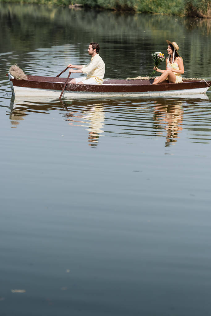 Seitenansicht von glücklicher Frau mit Blumen und romantischer Bootsfahrt mit Mann  - Foto, Bild