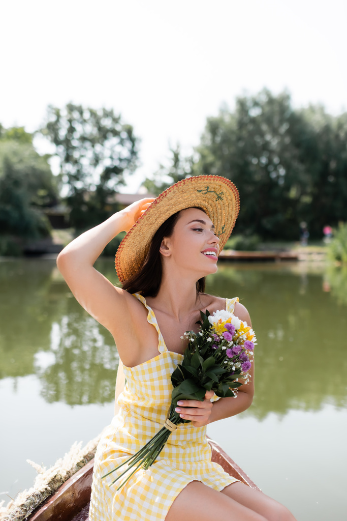 веселая молодая женщина в соломенной шляпе и платье с цветами во время прогулки на лодке по озеру - Фото, изображение