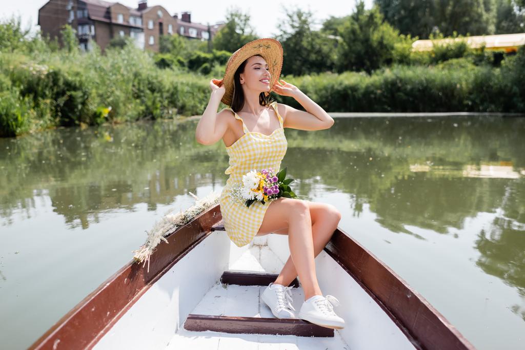 sorridente giovane donna regolazione cappello di paglia e avendo giro in barca sul lago - Foto, immagini