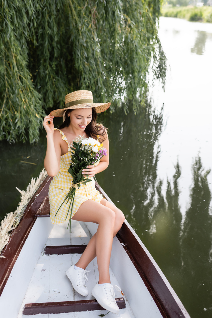 heureux jeune femme en chapeau de paille et robe tenant des fleurs pendant la promenade en bateau sur la rivière - Photo, image