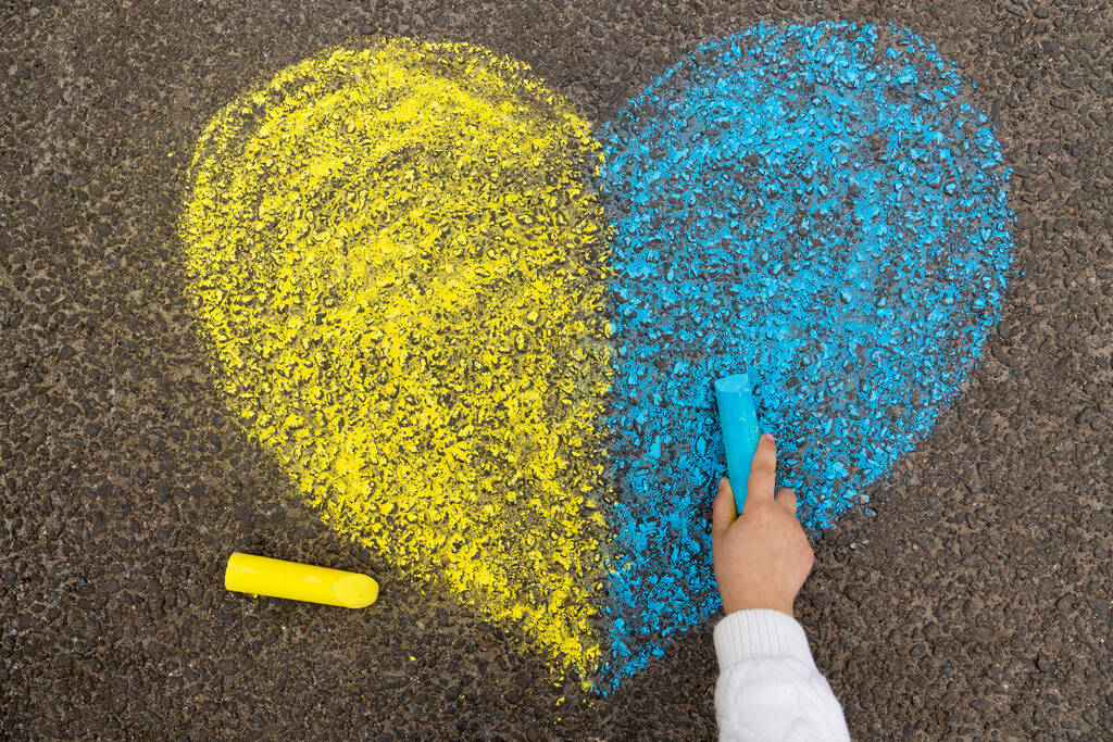 Прекратить войну на Украине. Маленькая девочка рисует сердце желтым и синим с мелом на тротуаре, как украинский флаг. Высокое качество фото - Фото, изображение