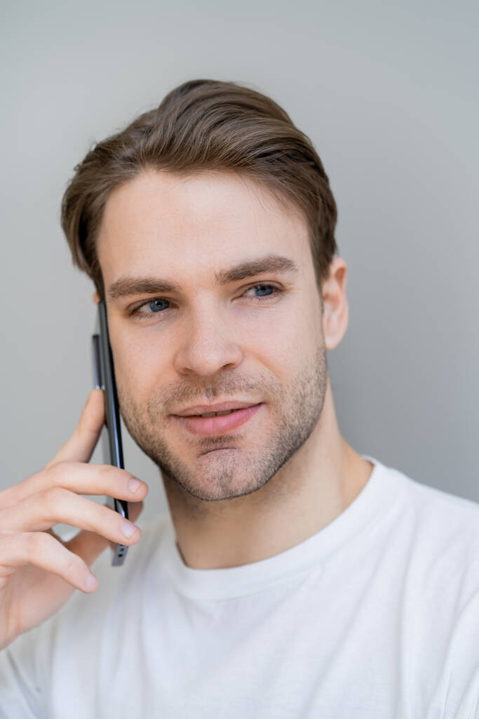 retrato del joven mirando hacia otro lado durante la conversación en el teléfono inteligente aislado en gris - Foto, Imagen