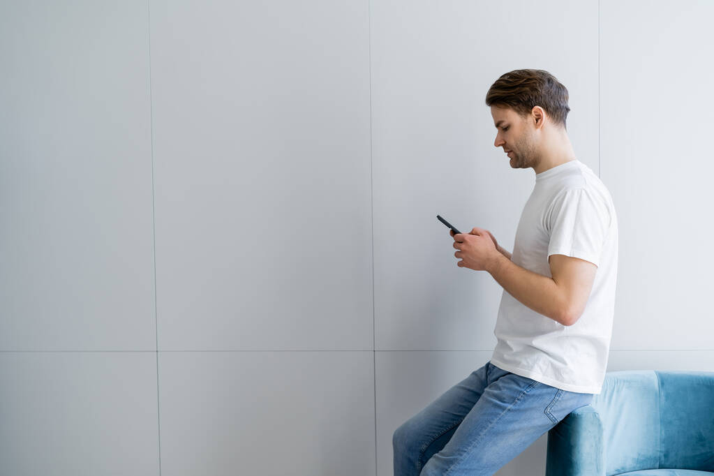 widok z boku młody człowiek w białej koszulce za pomocą telefonu komórkowego w pobliżu szarej ściany - Zdjęcie, obraz