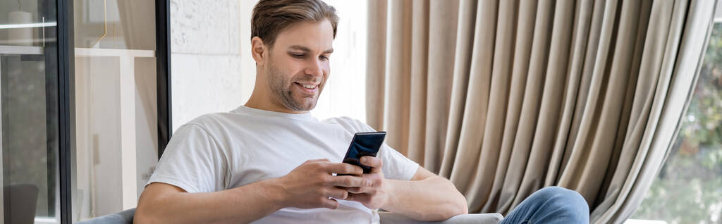 mensajería hombre feliz en el teléfono inteligente mientras está sentado en casa cerca de cortina beige, pancarta - Foto, Imagen
