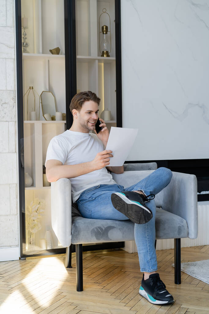 vue complète de l'homme positif avec du papier parlant sur un téléphone portable dans un fauteuil - Photo, image