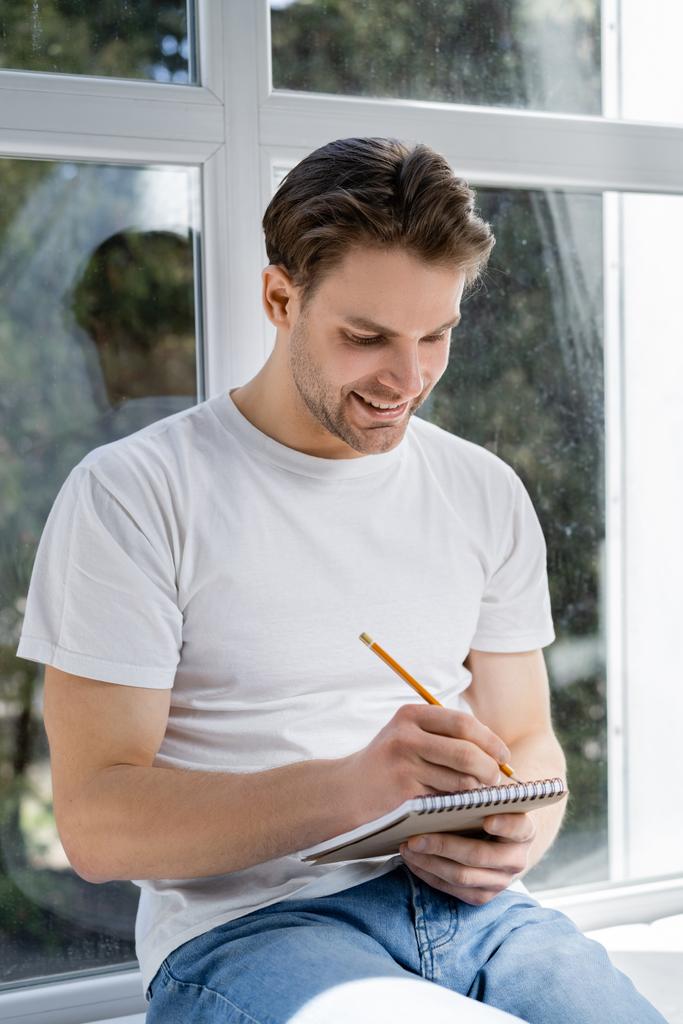Lächelnder Mann schreibt in Tagebuch in der Nähe von Fenster zu Hause - Foto, Bild
