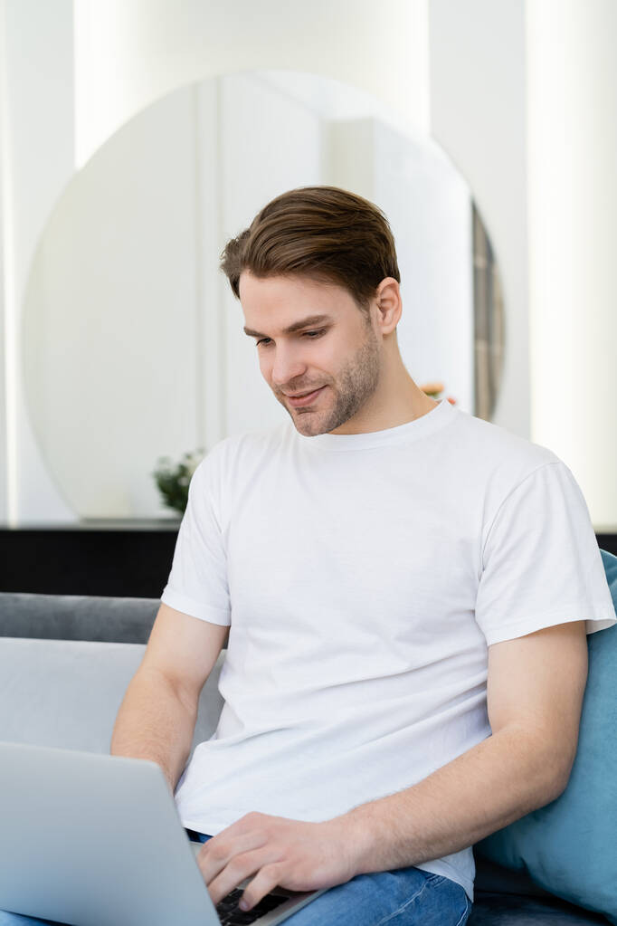young and smiling man in white t-shirt working on laptop at home - Photo, Image