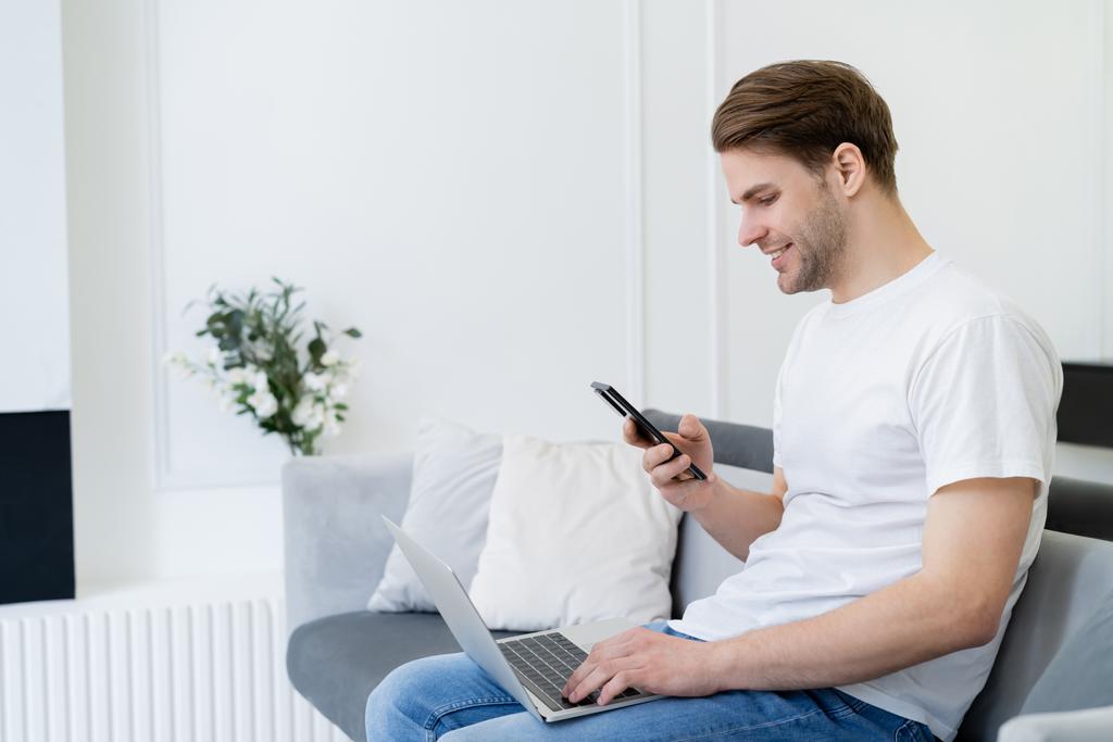 Seitenansicht eines lächelnden Mannes, der mit Smartphone und Laptop auf der heimischen Couch sitzt - Foto, Bild