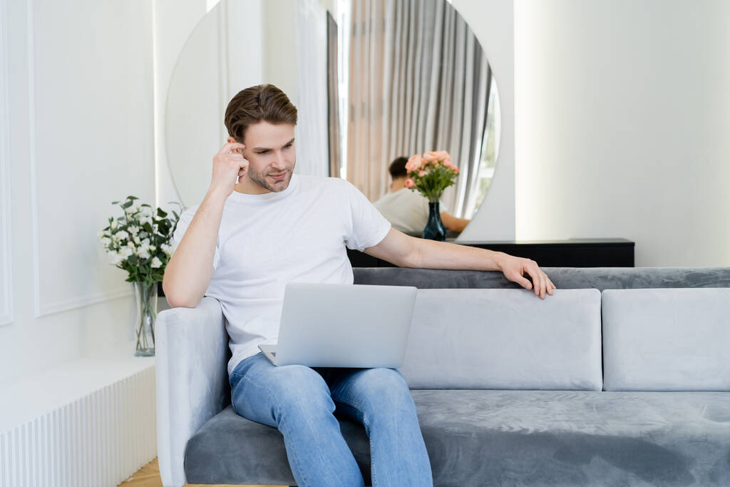 joven hombre pensativo sentado en el sofá cerca de jarrones con flores y mirando a la computadora portátil - Foto, imagen