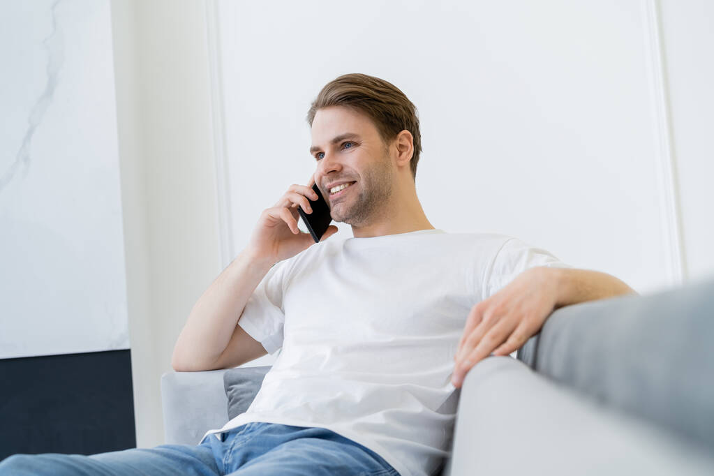 Χαρούμενος νεαρός που κάθεται σε θολό καναπέ και μιλάει στο κινητό τηλέφωνο - Φωτογραφία, εικόνα