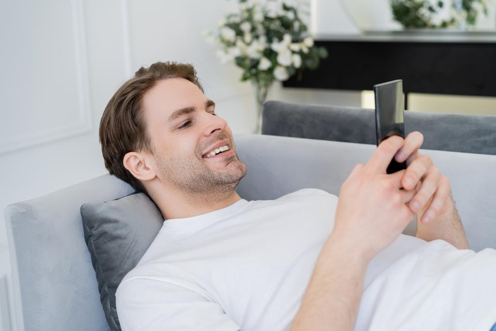 contento giovane uomo che riposa sul divano e chatta su smartphone - Foto, immagini