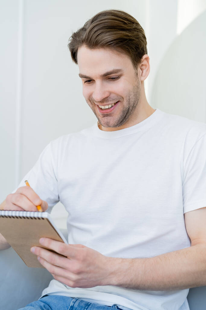 молодой и счастливый мужчина в белой футболке, пишущий в дневнике дома - Фото, изображение